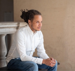 man sits with hands together in front of columned half wall